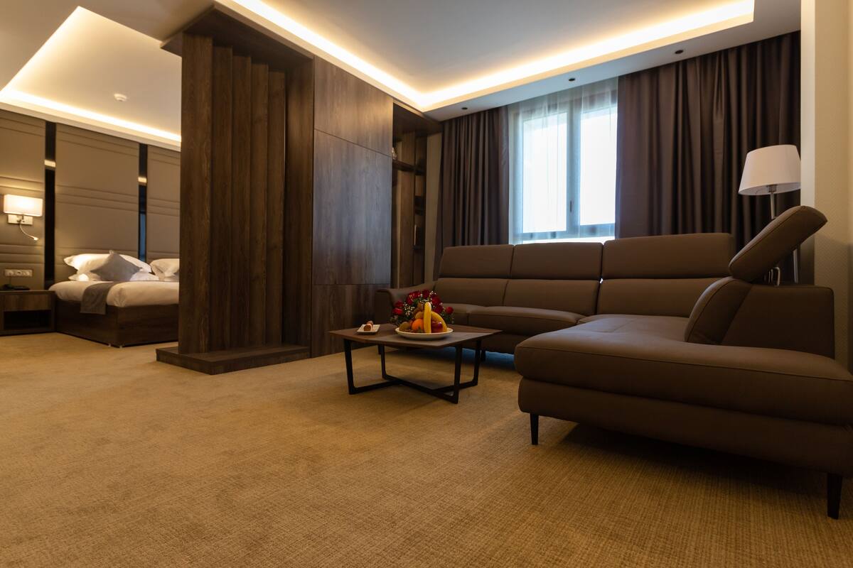 A modern hotel room with a sofa, a small table with fruit, a bed, and a large window with curtains.