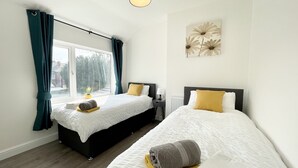 A hotel room with two beds, a window with curtains, a bedside table with a lamp, and a wall-mounted flower painting. - Worksop Newly Refurbished 2-bedroom House (Worksop)