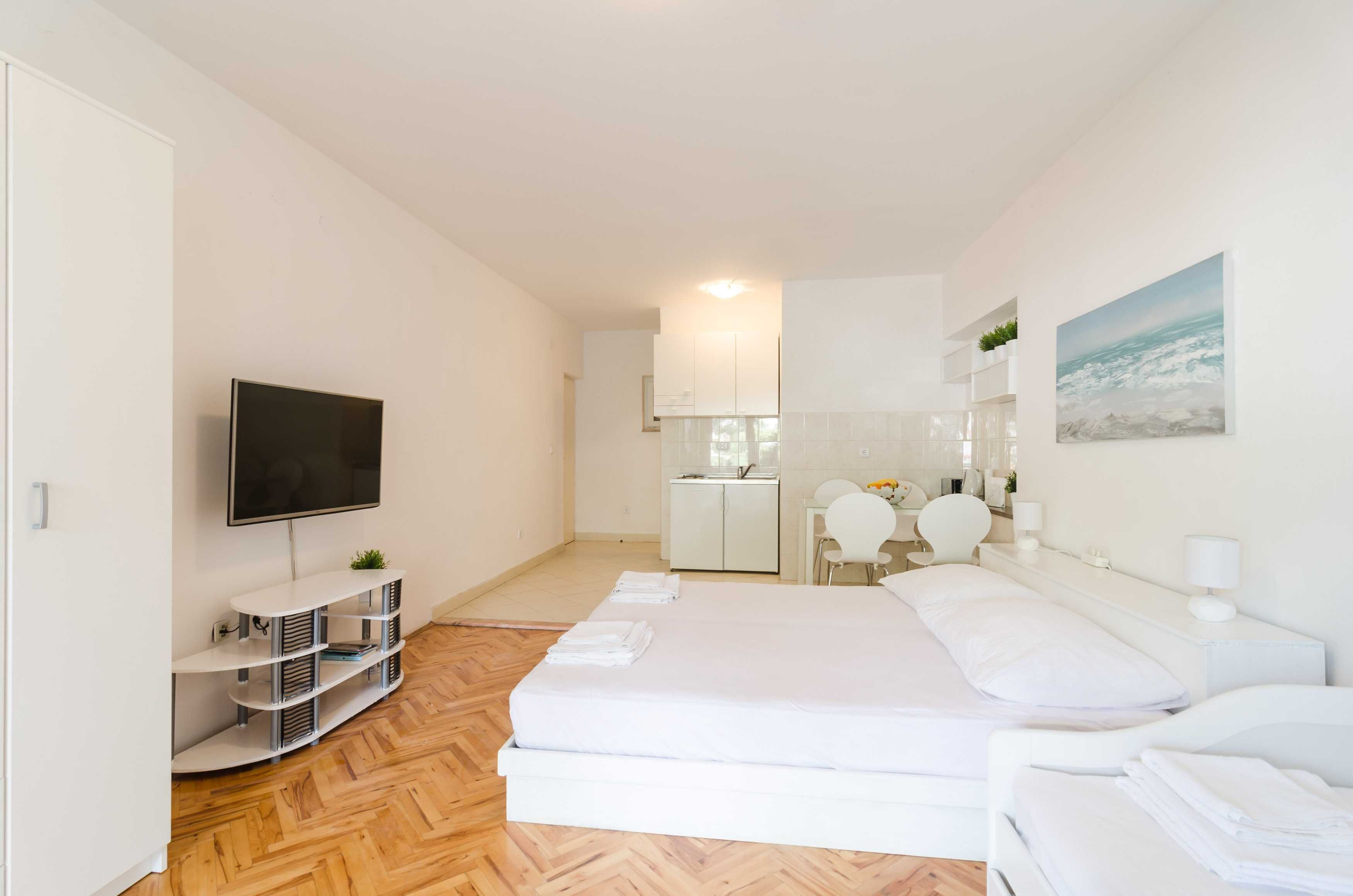 A modern hotel room with a large bed, a TV, a small kitchenette, and a seating area.