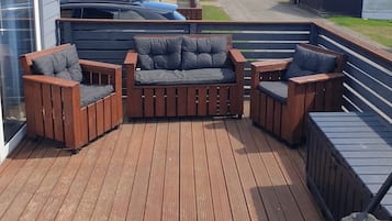 A wooden deck with a sofa and chairs, a glass door, and a view of a road and buildings.