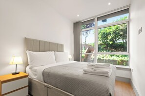 Un lit bien fait, recouvert d’une couverture grise, une table de chevet en bois avec une lampe, et une grande fenêtre donnant sur un paysage verdoyant.