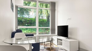 A modern room with a glass table, white chairs, a sofa, a TV on a white cabinet, and a large window with a view of greenery. - Charming 1-bed Apartment in Harrow (Harrow)