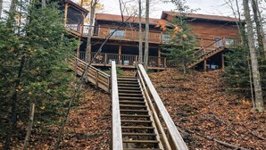 A wooden staircase leading to a cabin with a balcony, surrounded by trees and fallen leaves.
