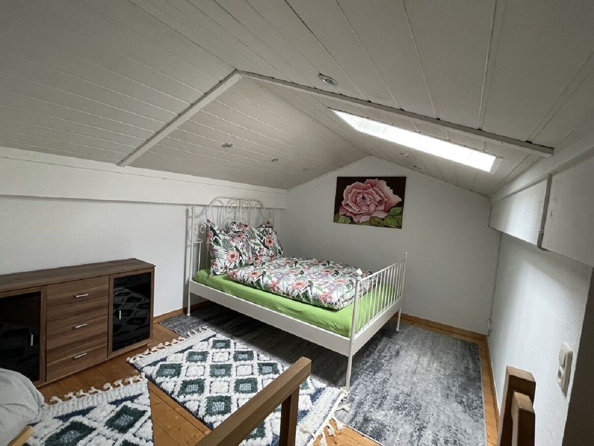 Un petit appartement d’une seule pièce, avec un plafond en pente, un lit recouvert d’une courtepointe à motifs floraux, une armoire en bois et un tapis à motifs.