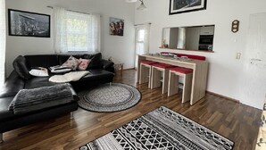 A living room with a black leather sofa, a wooden dining table with red chairs, a kitchen area with a microwave, and a large window with curtains. - 2 Bedroom Apartment in Kempten (Kempten)