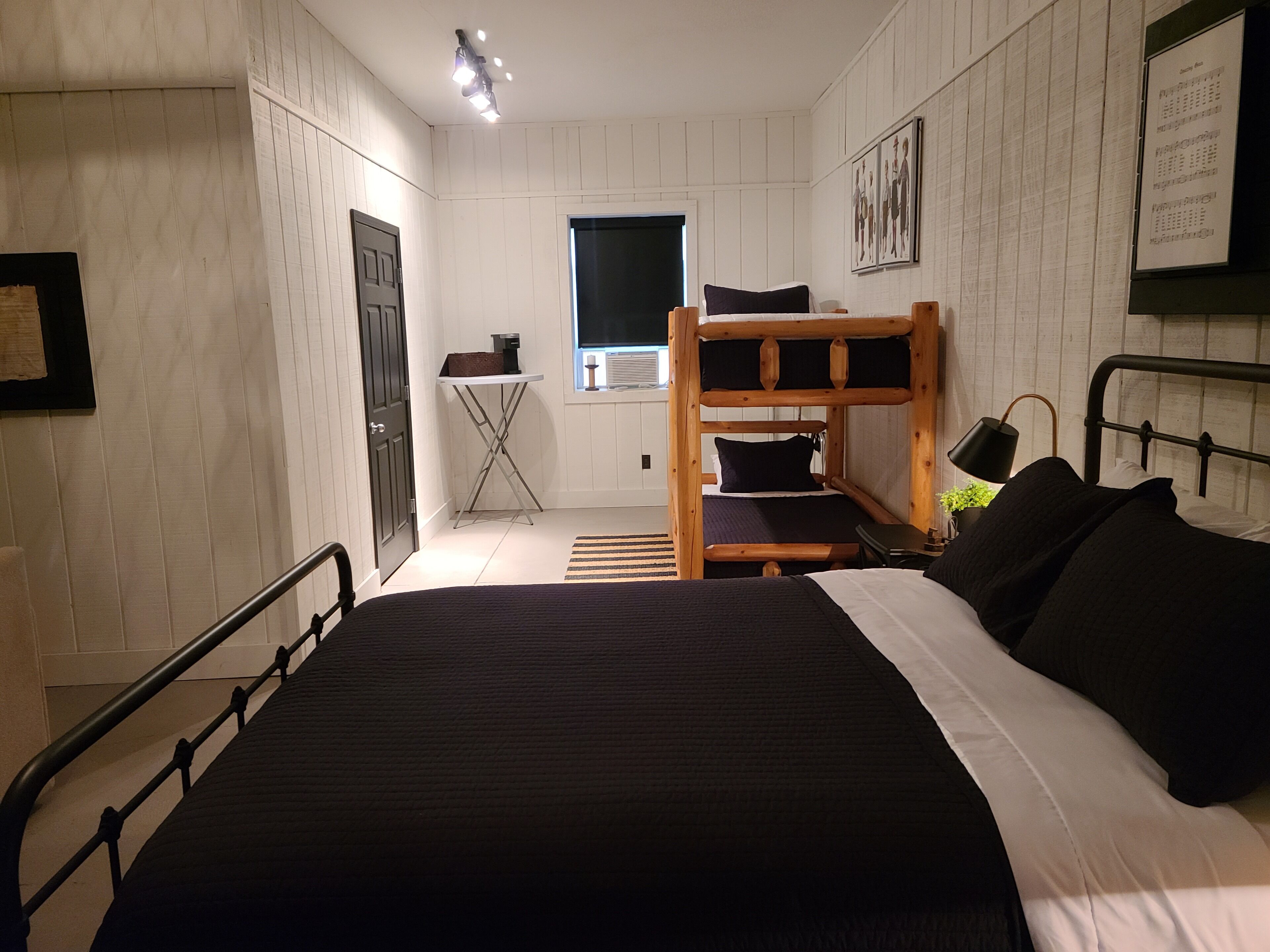 A bunk bed with a black mattress and pillows, a small table with a lamp, and a window with a view of the outside.