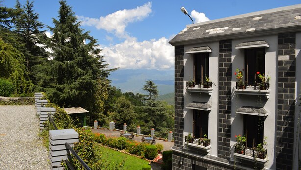 A building with a stone facade, multiple balconies with potted plants, and a clear view of the surrounding forest and mountains. - Sun N Snow Inn By Leisure Hotels (Somashwar)