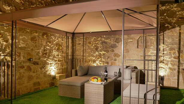 A cozy outdoor seating area with wicker furniture, a table with a fruit bowl and wine glasses, and a stone wall backdrop.