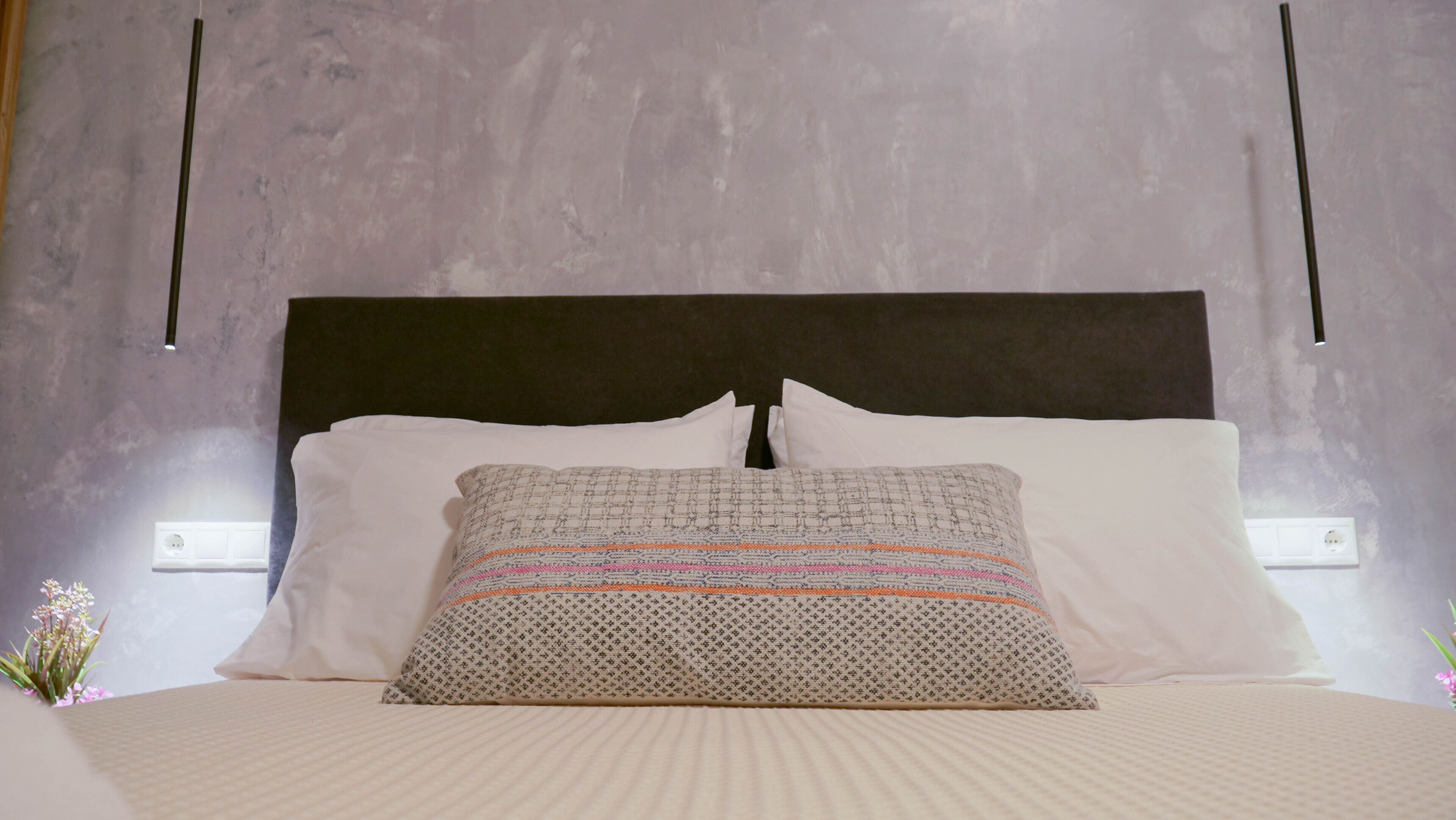 Una cama bien tendida con un cojín estampado y cojines blancos, un cabecero oscuro y una pared con textura y dos lámparas.