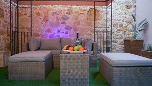 A cozy outdoor seating area with wicker furniture, a stone wall backdrop, and a table with fruits and a bottle.