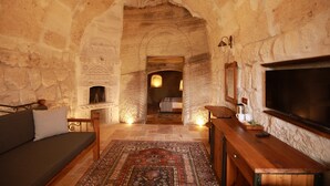 A cozy room with a stone fireplace, a sofa, a wooden table, and a TV. - Sarnich Cave Suites (Nevsehir)