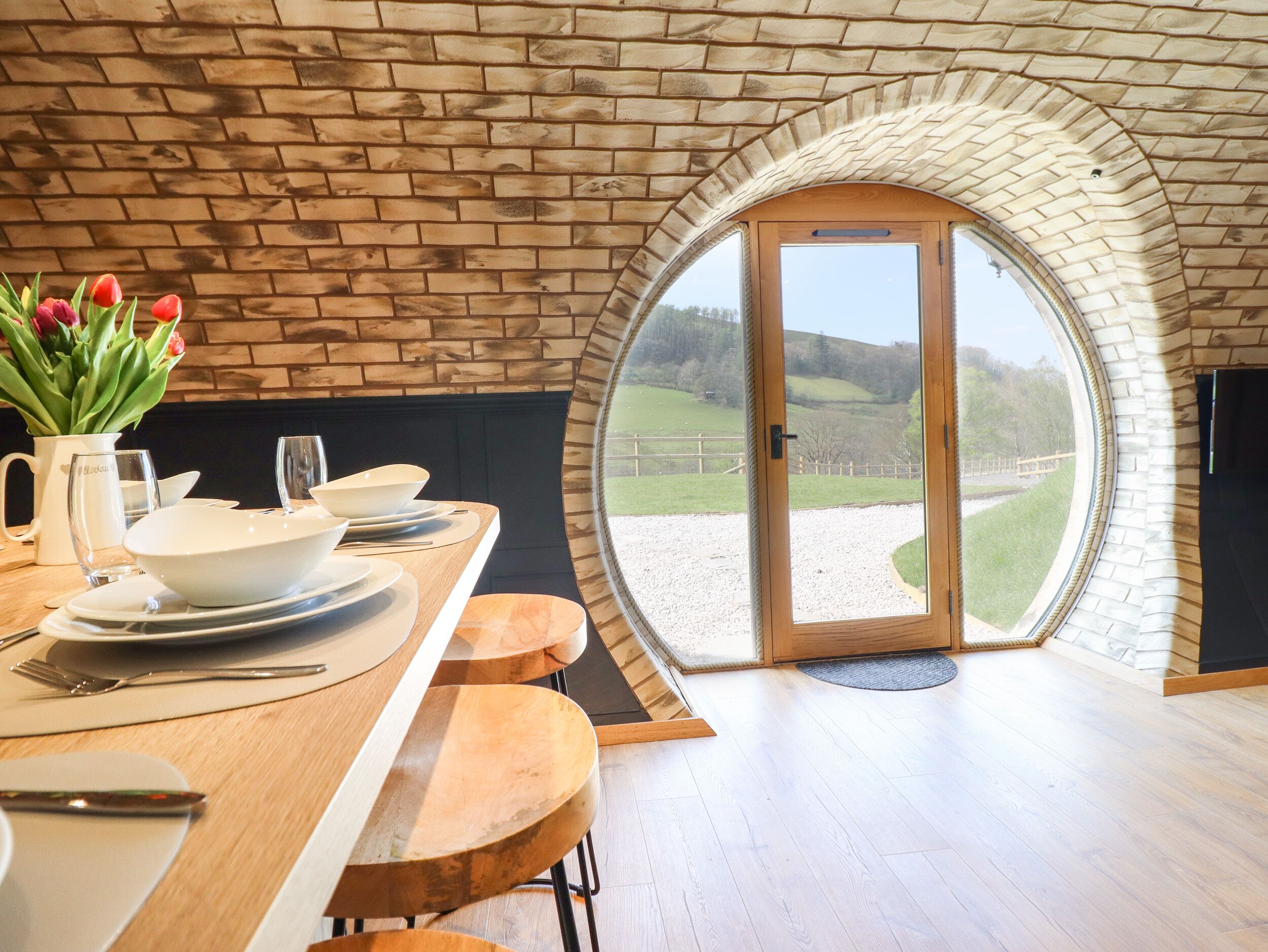 A dining area with a wooden table, white plates, and tulips. There is a large arched window with a view of a grassy landscape.