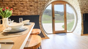 Ein Essbereich mit einem Holztisch, weißen Tellern und Tulpen. Ein großes, rundbogenförmiges Fenster bietet Blick auf eine grüne Landschaft.