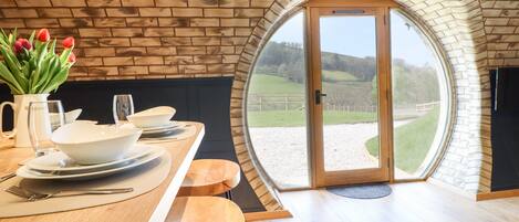 A dining area with a wooden table, white plates, and tulips. There is a large arched window with a view of a grassy landscape.