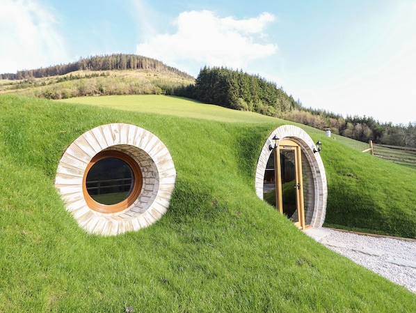 Two circular stone structures with wooden doors and windows, set on a grassy hill with a forested background.