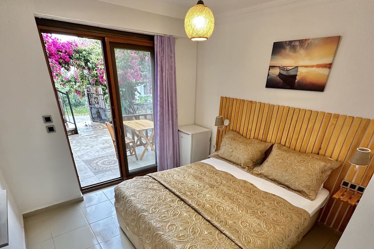 A bedroom with a bed, a small table, a chair, a ceiling light, and a view of the outside.