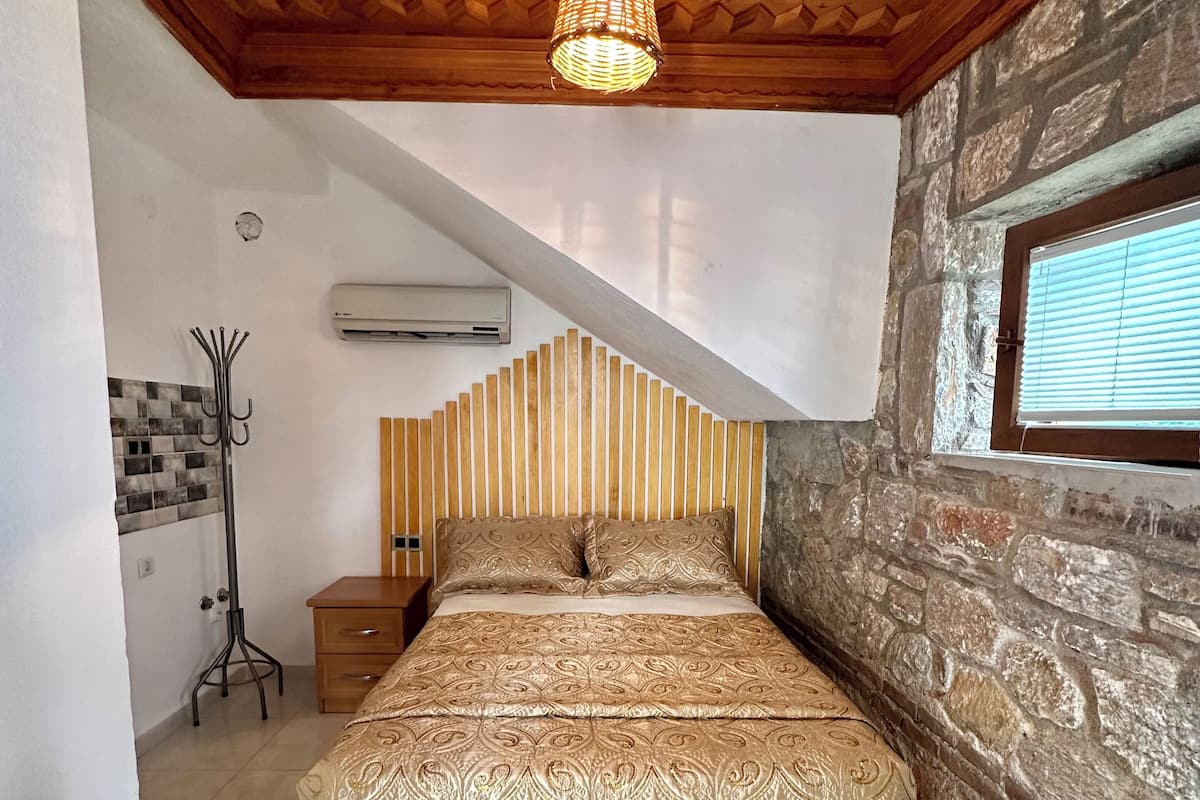 A bedroom with a bed, wooden ceiling, stone wall, and a window.