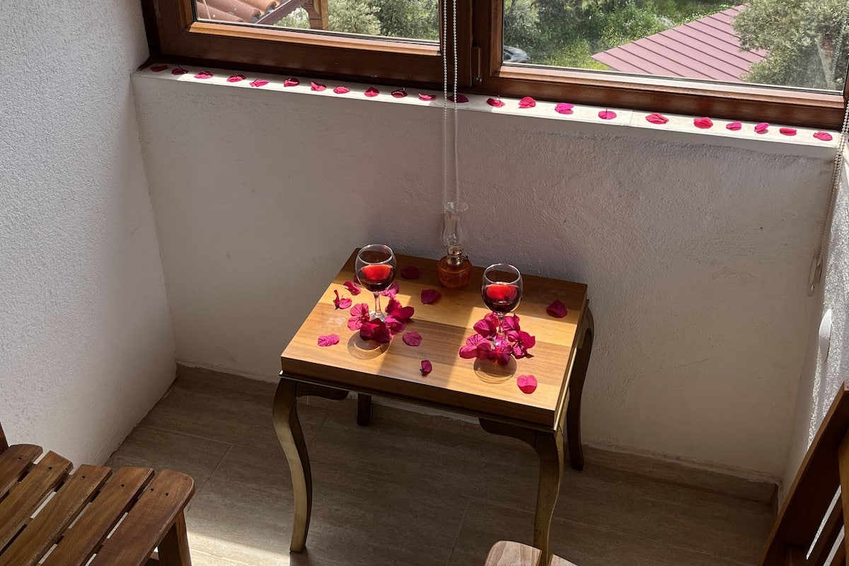 A room with a view of a lake and mountains, a wooden table with two glasses and rose petals, and a chair.