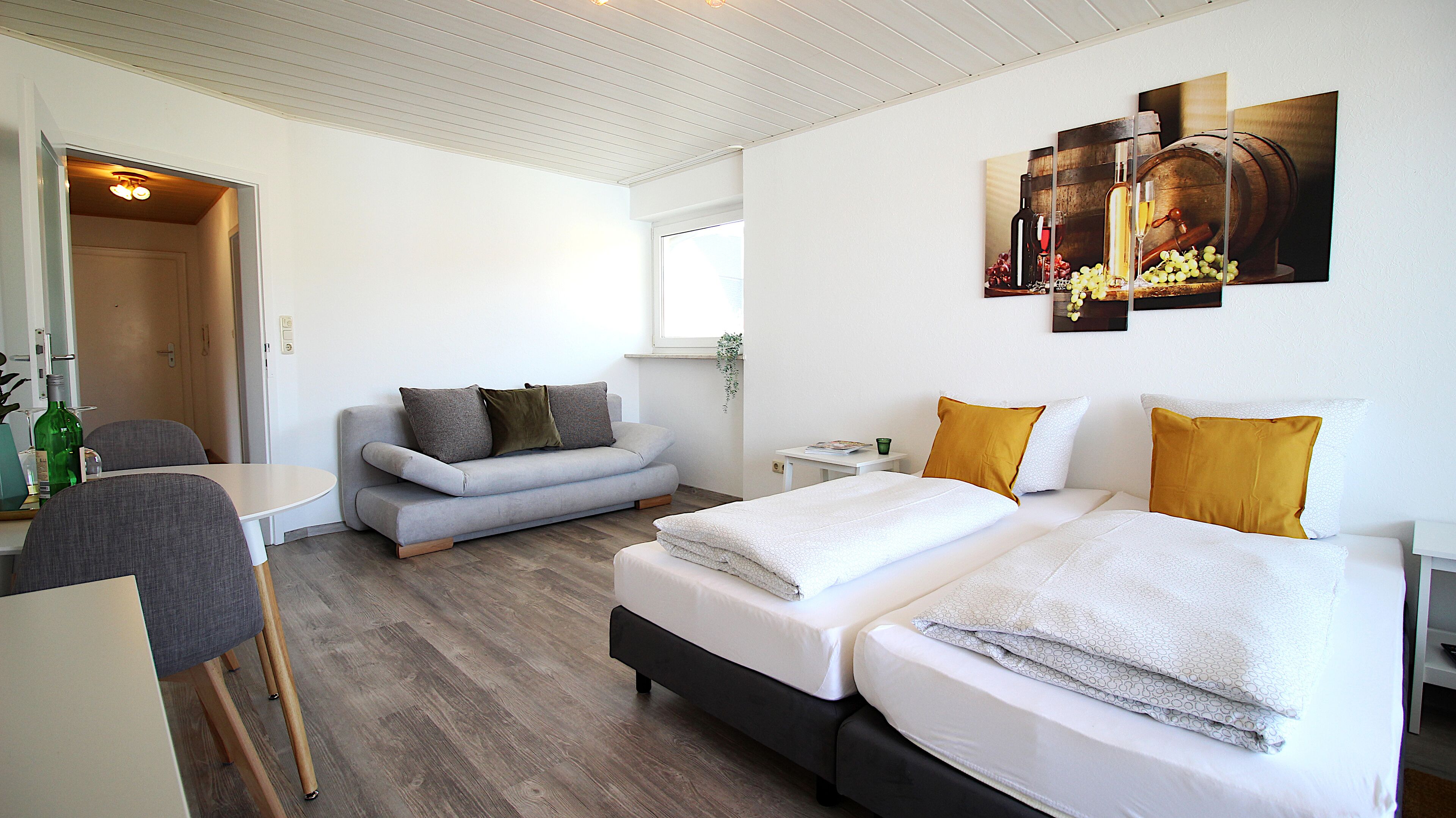 A modern hotel room with two beds, a sofa, a dining table, and a small kitchen area.