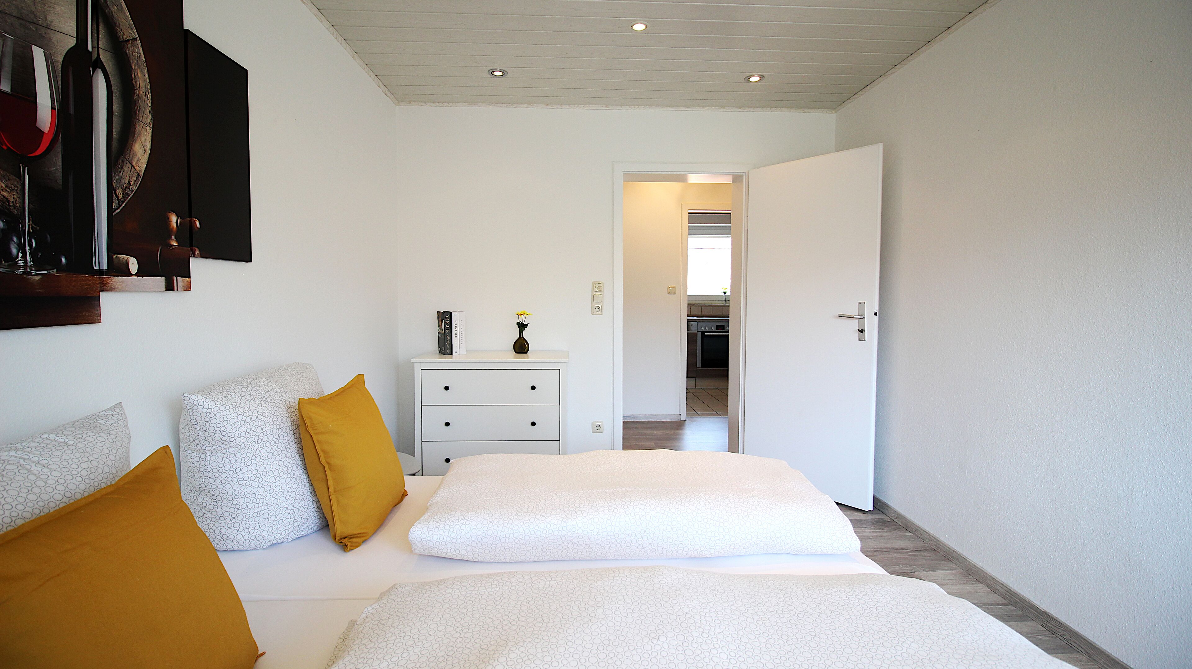 A bedroom with a bed, two pillows, a nightstand, and a door leading to a kitchen.