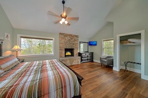 A bedroom with a bed, a stone fireplace, a television, and a window with a view of trees.