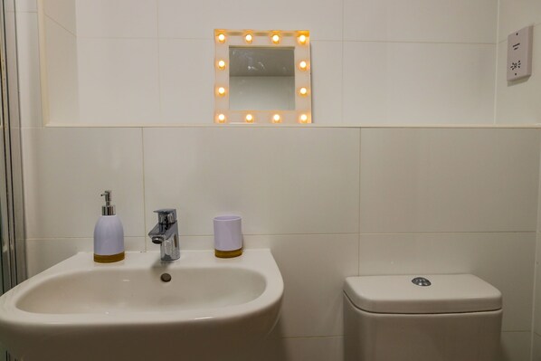 A bathroom with a sink, a mirror with lights, a toilet, and a soap dispenser.
