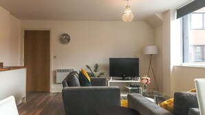 A modern living room with a flat-screen TV, grey sofas, a small round table, a standing lamp, and a large window.