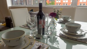 A dining table set with wine glasses, a bottle of wine, and bowls on a glass table with a view of buildings outside.