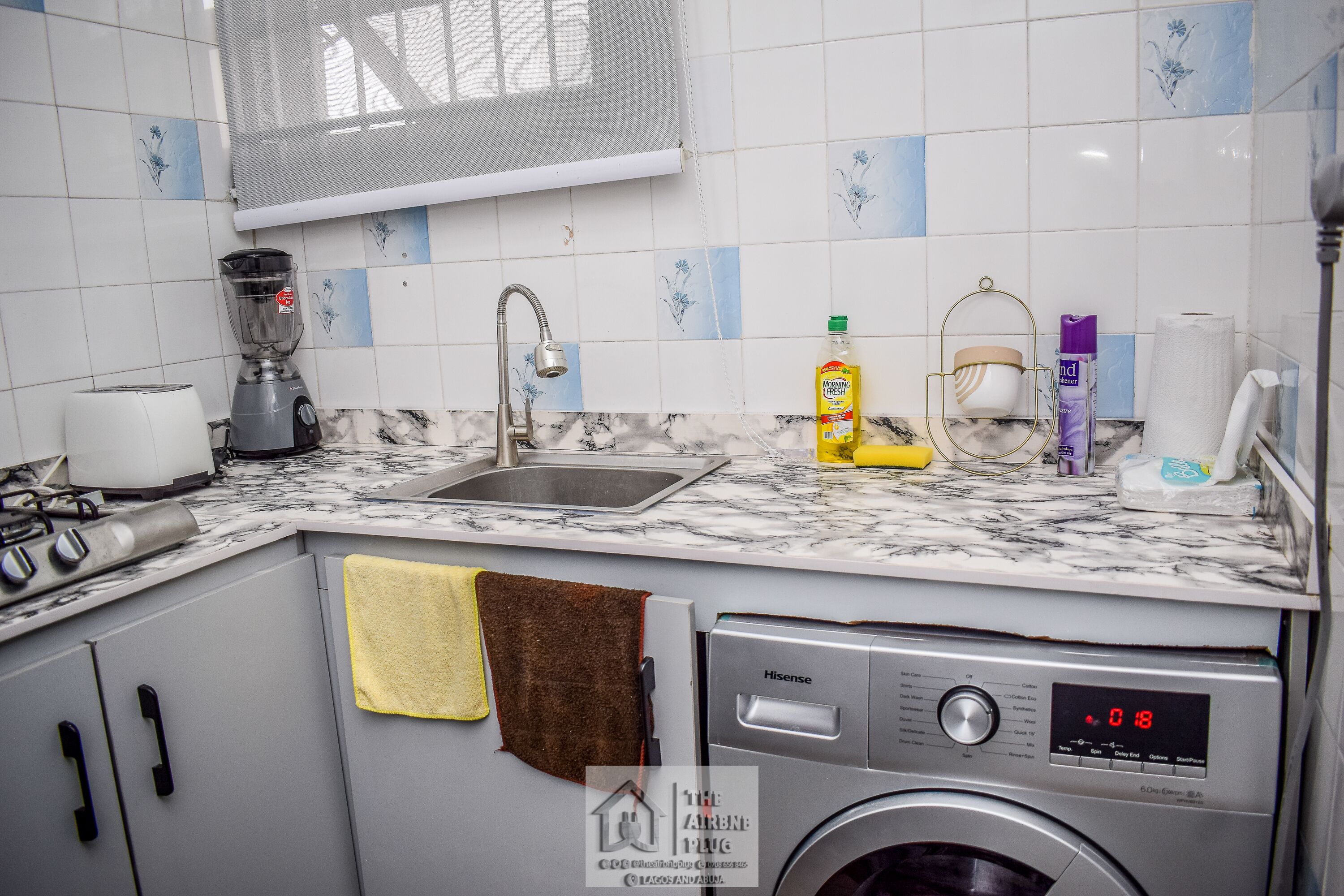 Une cuisine équipée d’un évier, d’un mixeur, d’un lave-linge et d’une fenêtre munie d’un moustiquaire.