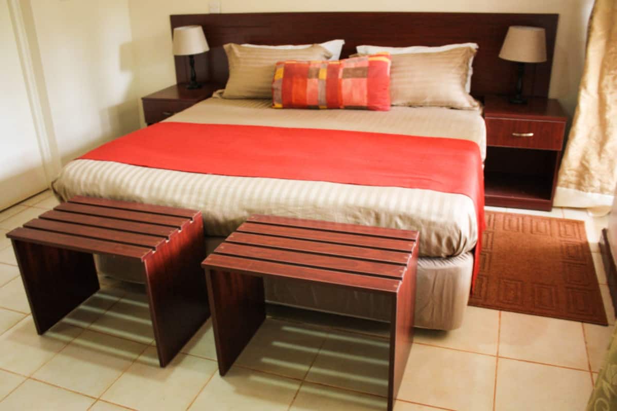 Habitación de hotel ordenada con una cama grande, dos mesitas de noche con lámparas, una colcha rojo y beige, y dos mesitas de madera.