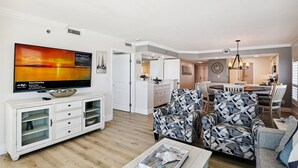 A modern living room with a flat-screen TV, a dining area with a chandelier, and a kitchen in the background. - Sea Oats 606 is a Gorgeous Gulf Front 3 BR - with Free Beach Service for 4 by RedAwning (Fort Walton Beach)