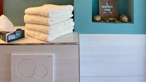 Um banheiro com parede azul-turquesa, um letreiro decorativo com a inscrição 'oh hello', uma pilha de toalhas brancas e objetos decorativos, incluindo uma âncora e uma estrela-do-mar.