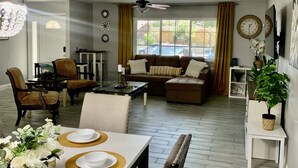 A dining area with a table set for four, surrounded by chairs. There is a sofa set in the background, a ceiling fan, and a window with curtains. - Cozy, Licensed home, 15min from the strip. (Las Vegas)