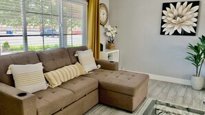 A living room with a sectional sofa, a clock, a flower painting, and a potted plant. - Cozy, Licensed home, 15min from the strip. (Las Vegas)