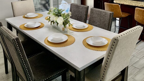A modern dining area with a white table set for six, surrounded by dark wooden chairs. A crystal chandelier hangs above, and a potted plant is on the table. - Cozy, Licensed home, 15min from the strip. (Las Vegas)