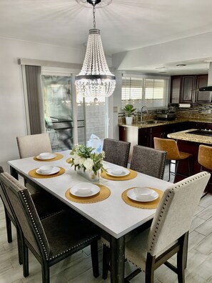 A modern dining area with a white table set for six, surrounded by dark wooden chairs. A crystal chandelier hangs above, and a potted plant is on the table. - Cozy, Licensed home, 15min from the strip. (Las Vegas)