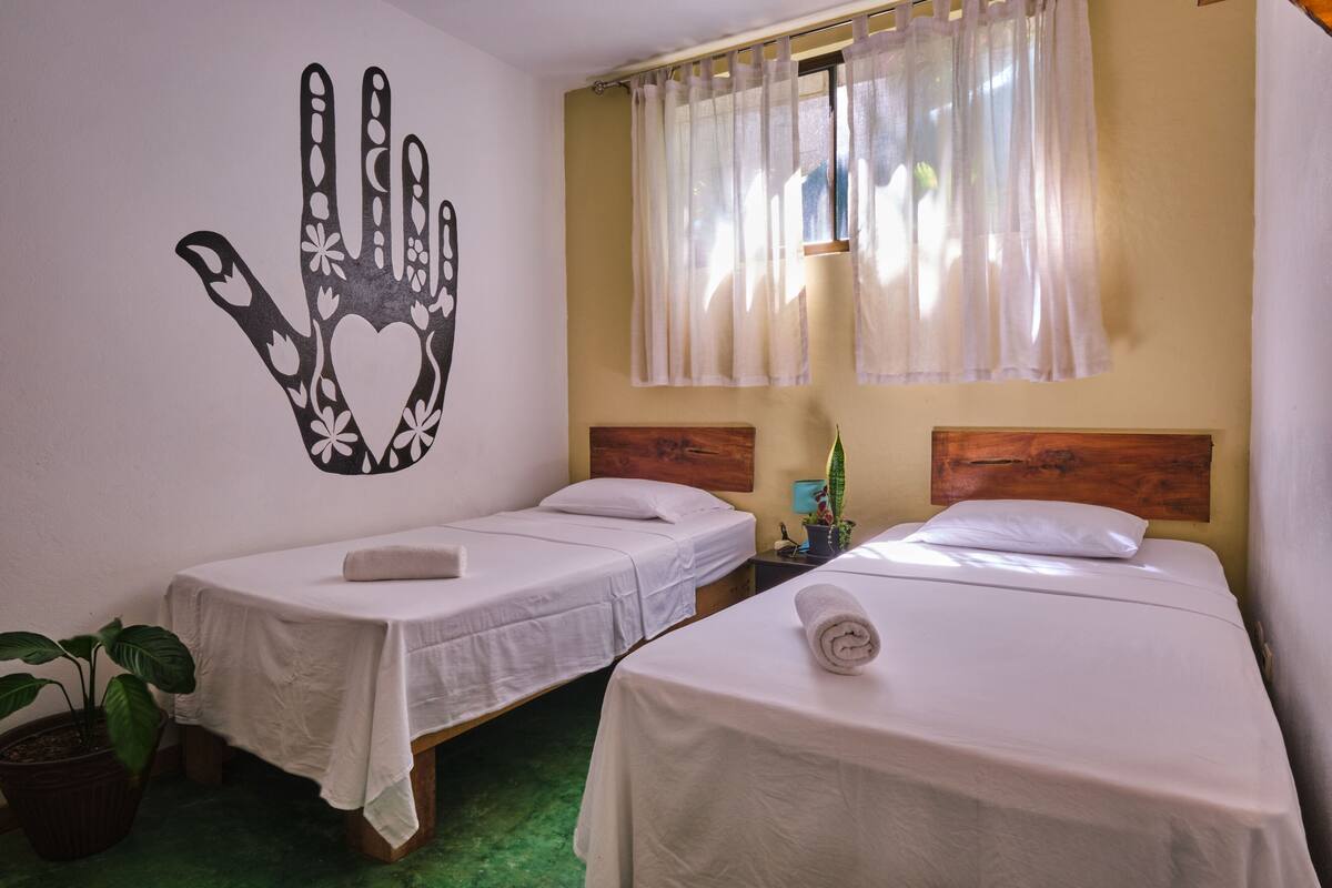 A room with two single beds, a hand-shaped wall decoration, a window with sheer curtains, and a potted plant.