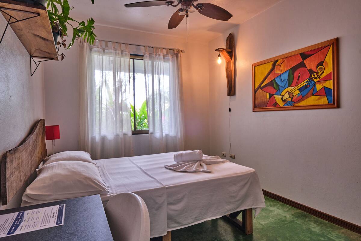 A hotel room with a bed, a desk, a ceiling fan, a window with curtains, a painting on the wall, and a plant hanging from the ceiling.