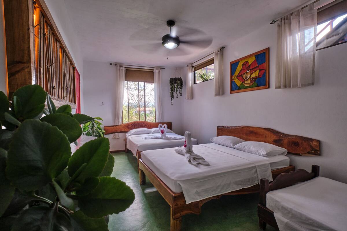 A hotel room with three wooden beds, a ceiling fan, a window with curtains, and a colorful painting on the wall.