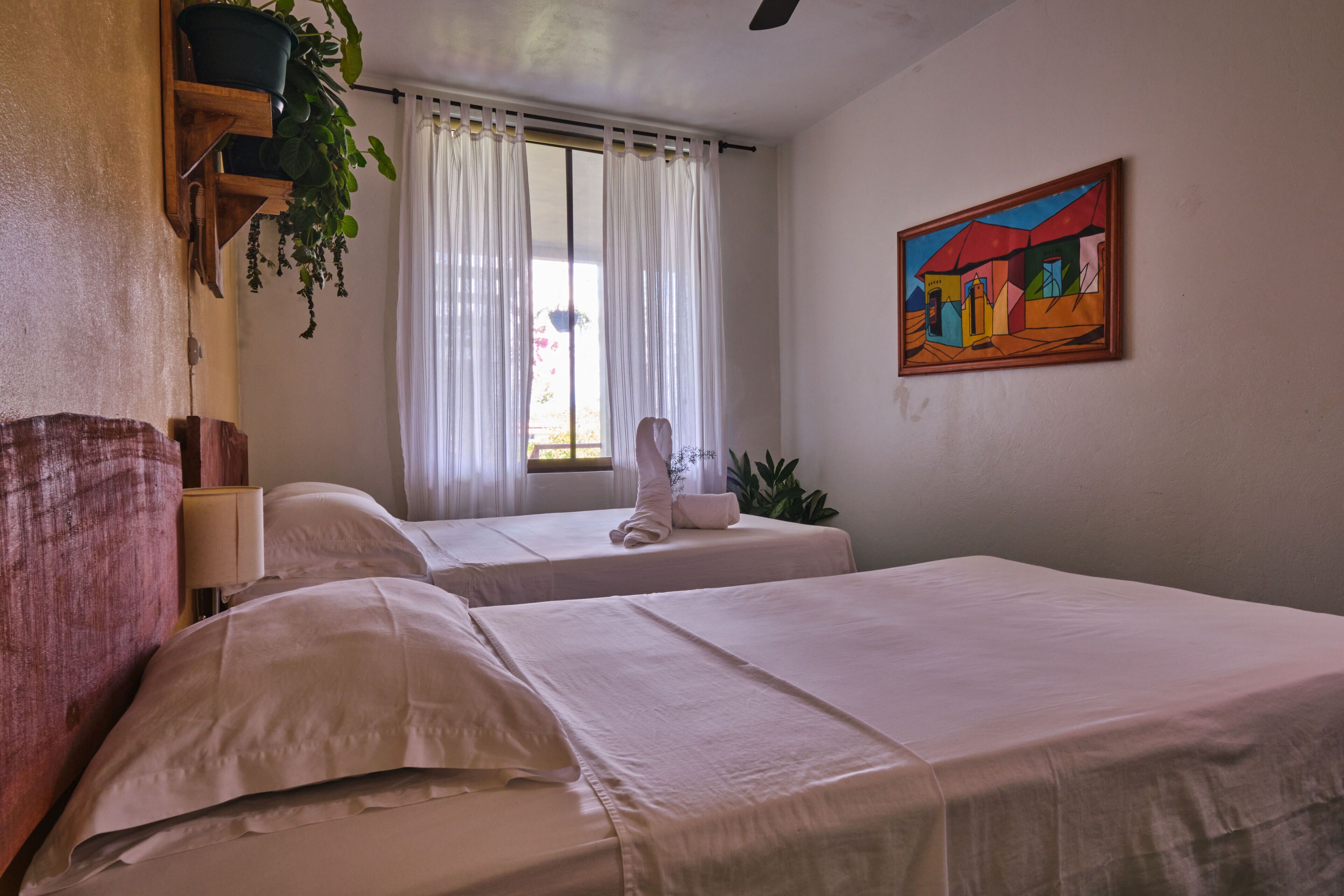 A hotel room with two beds, a window with white curtains, a painting on the wall, and a potted plant.