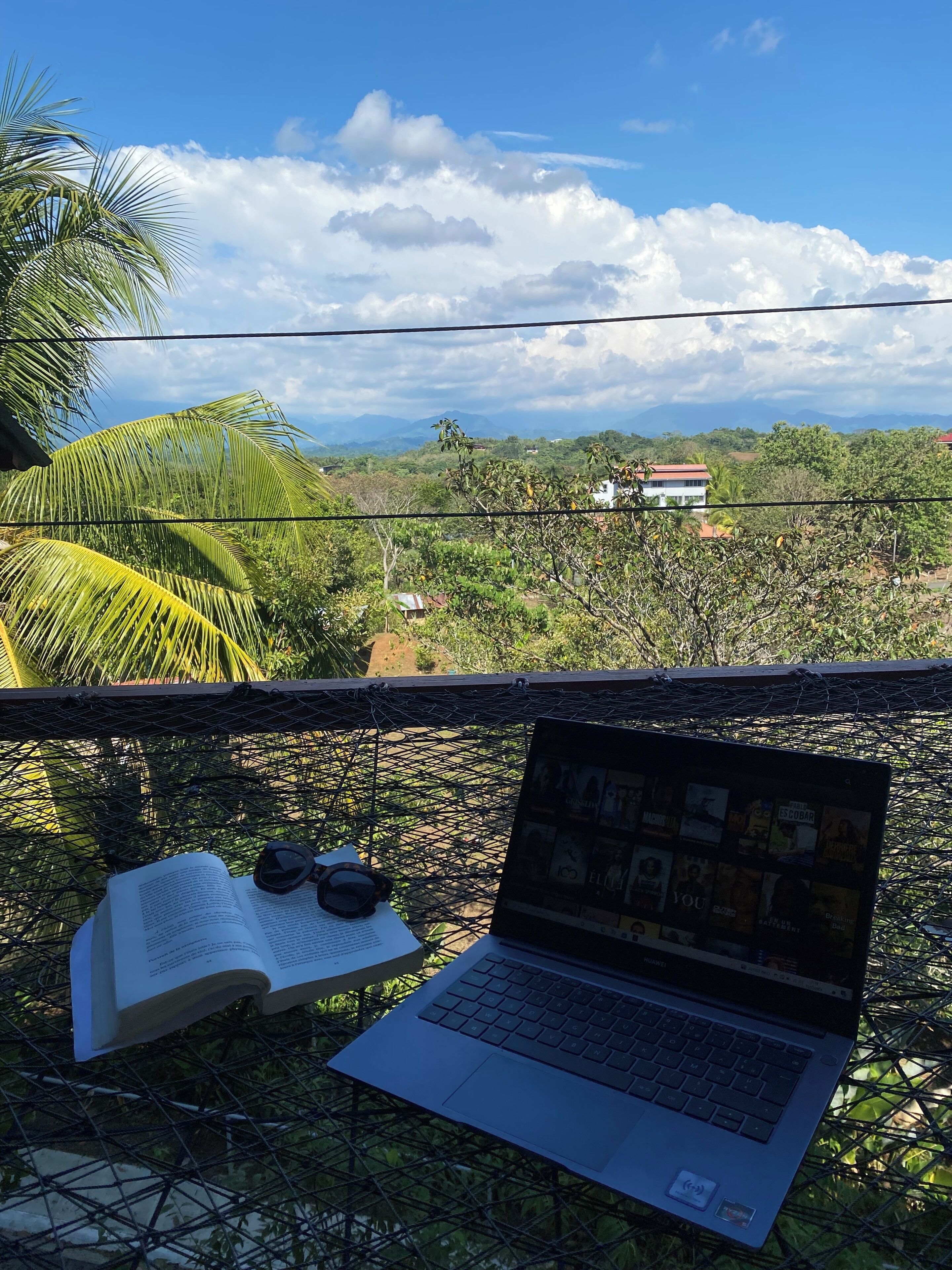 A laptop on a wire mesh table with an open book and sunglasses beside it, overlooking a lush landscape with houses and mountains.