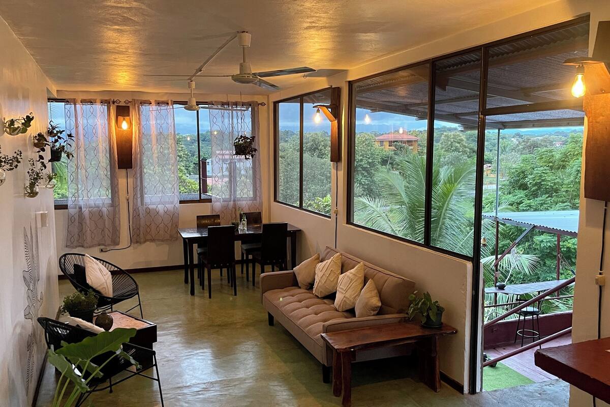 A cozy living area with a sofa, dining table, and chairs, large windows with curtains, and ceiling fans.