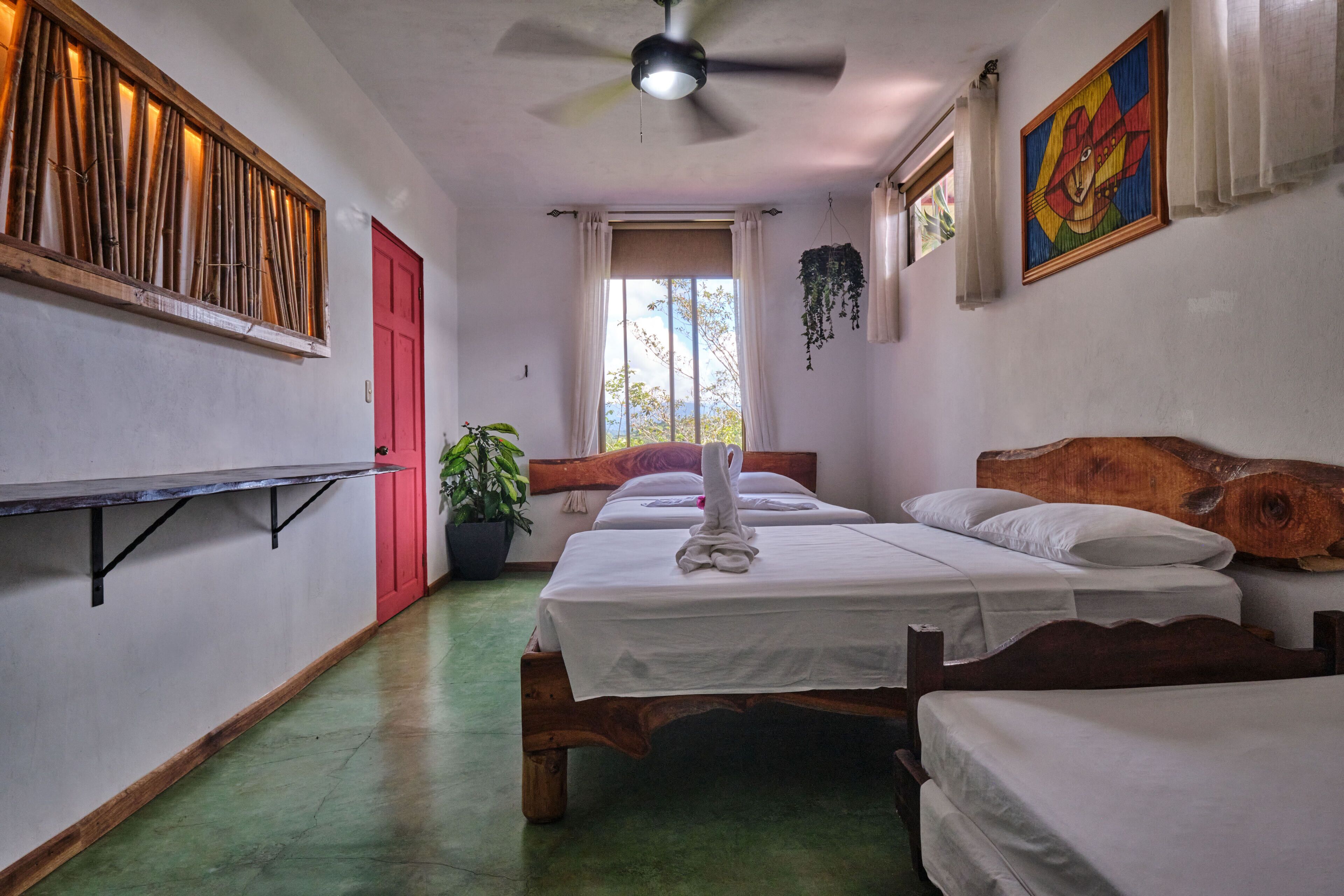 A room with two wooden beds, a ceiling fan, a red door, a window with curtains, and a wall-mounted shelf with plants.
