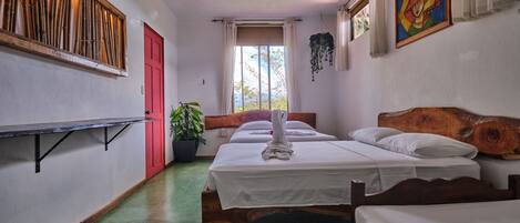 A room with two wooden beds, a ceiling fan, a red door, a window with curtains, and a wall-mounted shelf with plants.