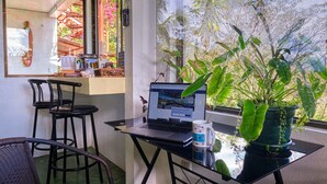 A workspace with a laptop, a coffee mug, and a potted plant on a balcony with a view of greenery and a building. - Villas Jacquelina (Quepos)