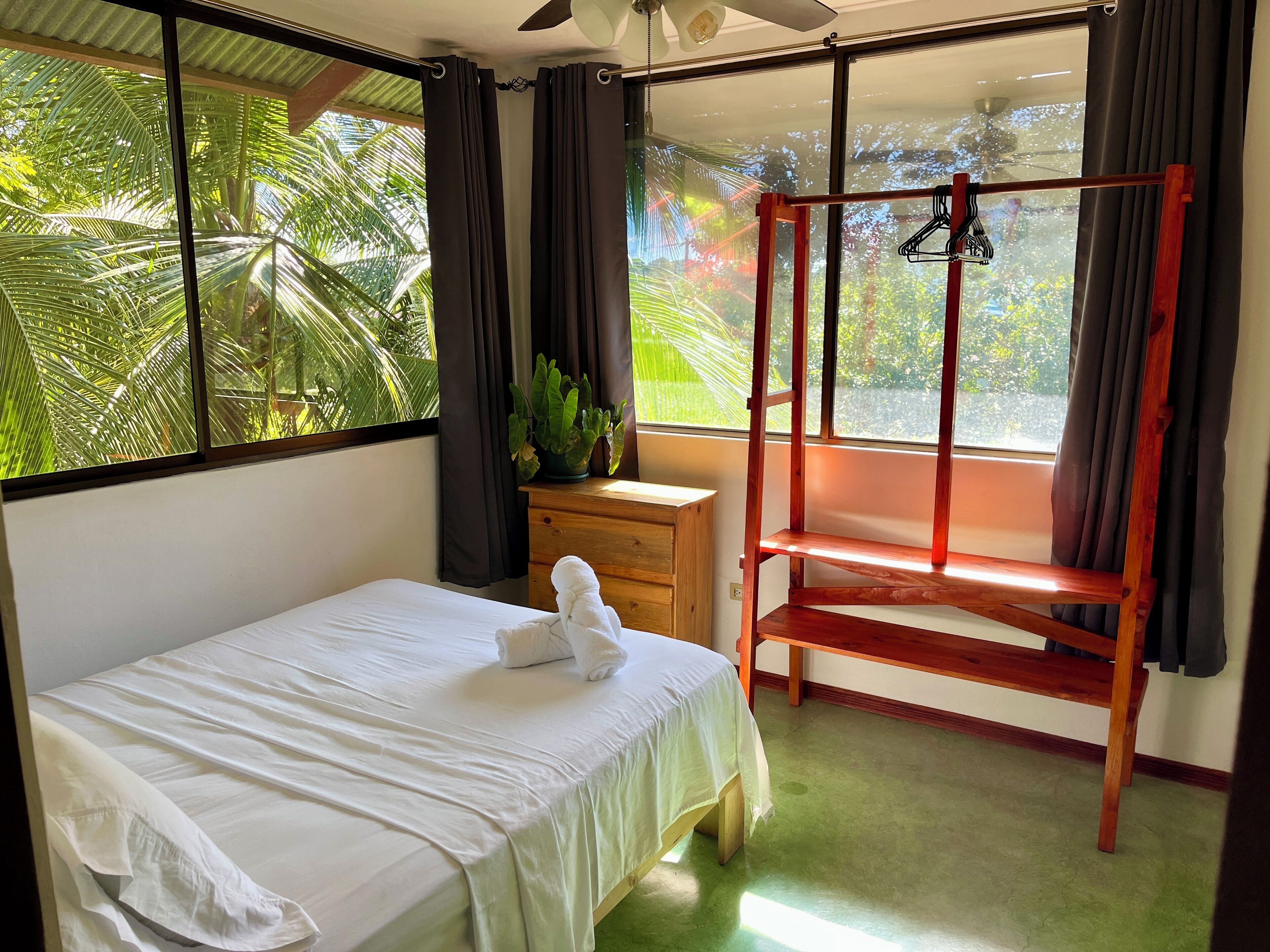 A bedroom with a bed, a wooden nightstand, a hanging chair, and a view of palm trees through the window.
