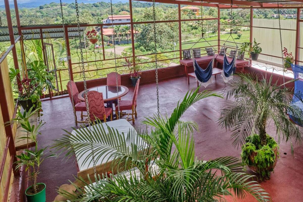 A covered outdoor area with a swing chair, a table, and chairs, surrounded by potted plants and a view of the countryside.