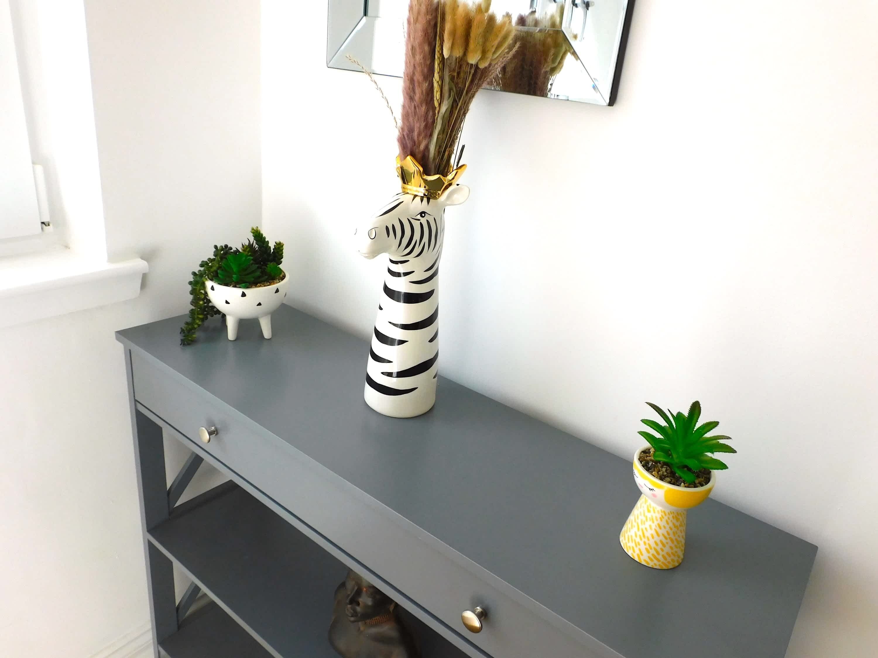 Ein grauer Konsolentisch mit einer Vase mit ZEBRA-MUSTER, einer kleinen Topfpflanze und einem dekorativen Gegenstand mit Federn.