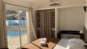 A hotel room with a bed, a desk, a chair, a window with curtains, an air conditioning unit, and a view of a swimming pool.
