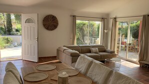 A spacious living room with a large sofa, a dining table with placemats, and a view of the garden through the sliding glass doors.